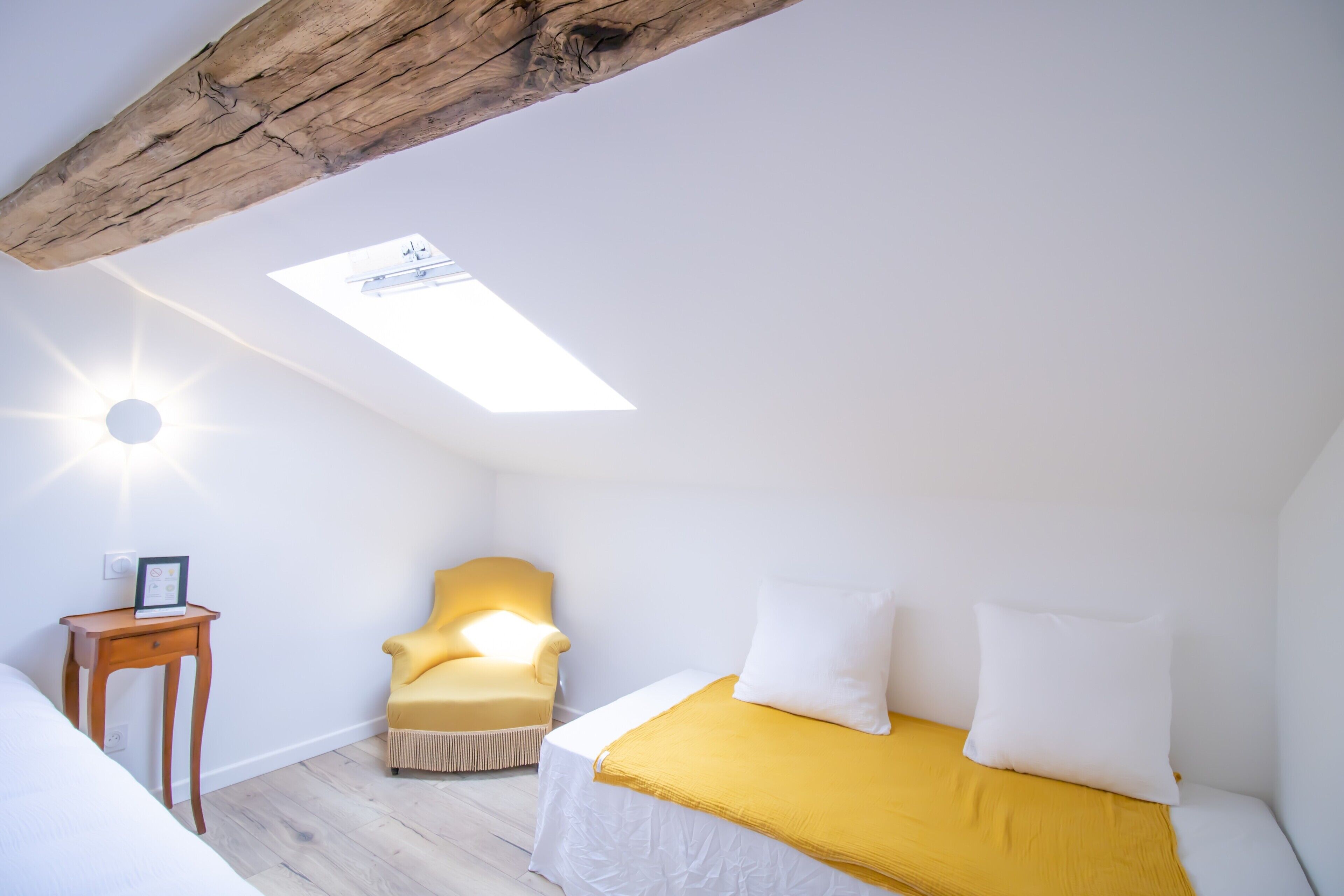 Une pièce avec un lit, un fauteuil jaune, une petite table, une lucarne et une poutre blanche au plafond.