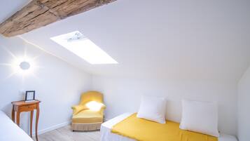 Une pièce avec un lit, un fauteuil jaune, une petite table, une lucarne et une poutre blanche au plafond.