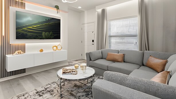 A modern living room with a large flat-screen TV mounted on a white wall, a grey sofa with brown cushions, a round coffee table with decorative items, and a window with blinds.
