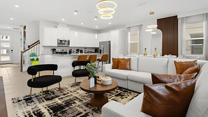 A modern living room with a white sofa, brown leather cushions, a coffee table, and black chairs. There is a kitchen area with white cabinets and a microwave. - Modern 6-BR Home w/ Summer Kitchen - Solara Resort (Kissimmee)