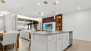 A modern kitchen with a central island, white cabinetry, and a built-in TV. - Modern 6-BR Home w/ Summer Kitchen - Solara Resort (Kissimmee)
