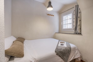 Uma cama de solte com roupa de cama branca e castanha, um candeeiro pendente, uma janela com cortinas e uma pequena prateleira com um objeto decorativo.