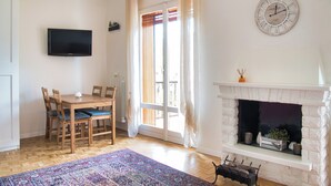 A living room with a wooden dining table, chairs, a flat-screen TV, a fireplace, and a large window with curtains. - Cableway to heaven - Studio Apartment, Sleeps 3 (Barzio)