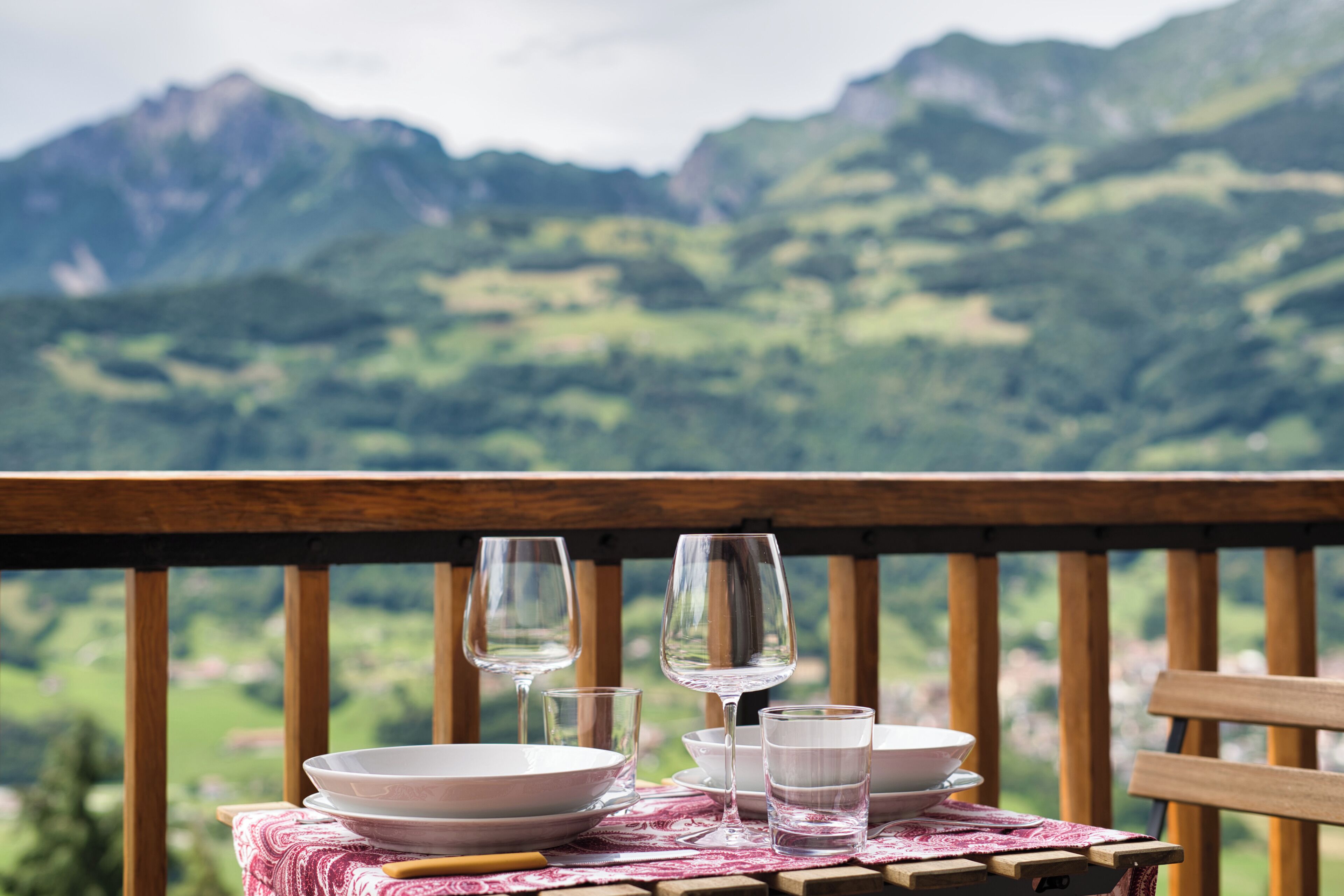Une table dressée avec des assiettes, des verres et un saladier, placée sur un balcon offrant une vue sur les montagnes.