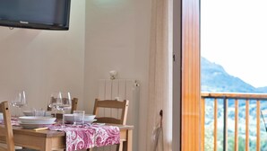 A dining table set with plates, glasses, and a red tablecloth. A television is mounted on the wall above the table. The room has a view of mountains through a balcony with a wooden railing. - Cableway to heaven - Studio Apartment, Sleeps 3 (Barzio)