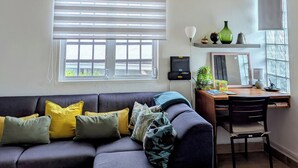 Una sala de estar con sofá, escritorio, silla, lámpara colgante y ventana con persianas.