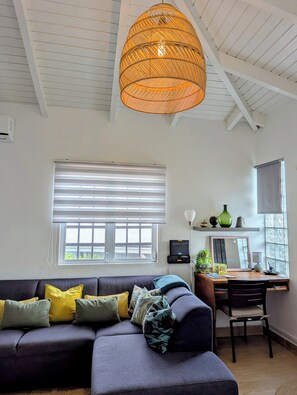 A living room with a sofa, a desk, a chair, a hanging light fixture, and a window with blinds.