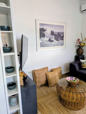 Una sala de estar con una televisión de pantalla plana, un cuadro enmarcado de un paisaje, una mesa de centro de mimbre y un aparador con cuencos decorativos.