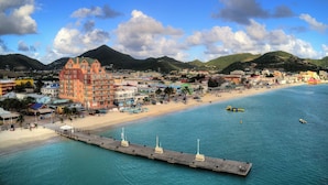 Un pueblo costero con un muelle que se adentra en el mar, edificios de colores y montañas al fondo.