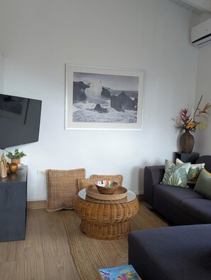 A living room with a flat-screen TV, a framed photograph of a coastal scene, wicker furniture, and a dark-colored sofa.
