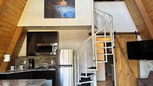 A modern kitchen with a stainless steel refrigerator, a built-in oven, and a coffee machine. There is a spiral staircase leading to an upper level.