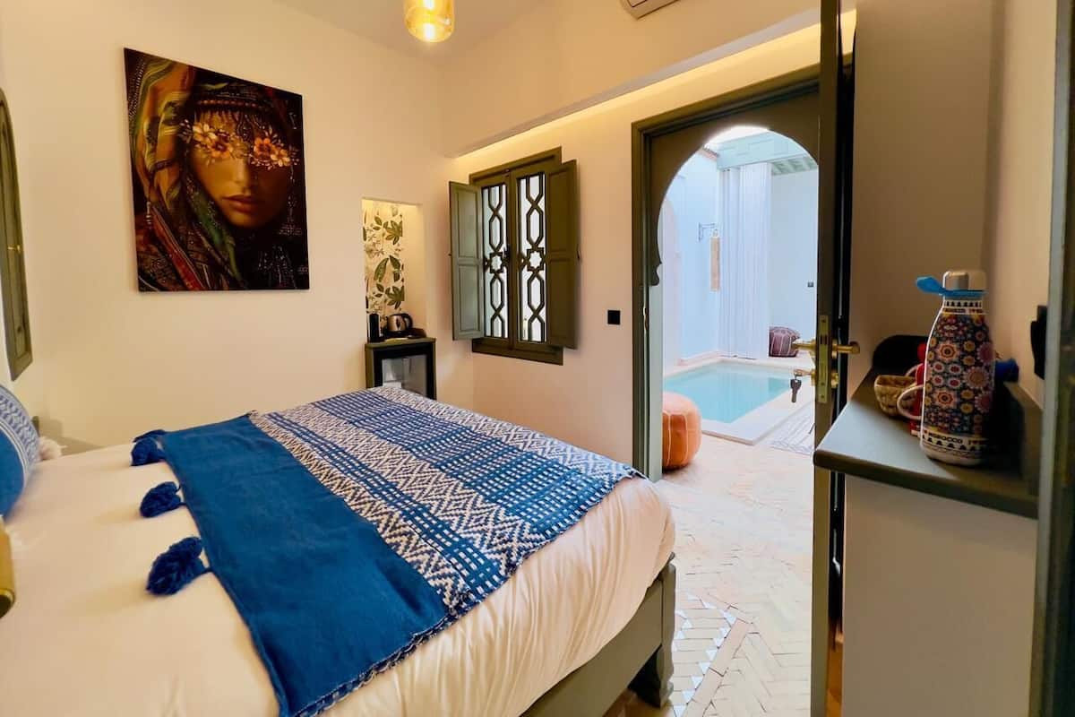 A bedroom with a bed, a painting, a window with shutters, and a view of an outdoor swimming pool.