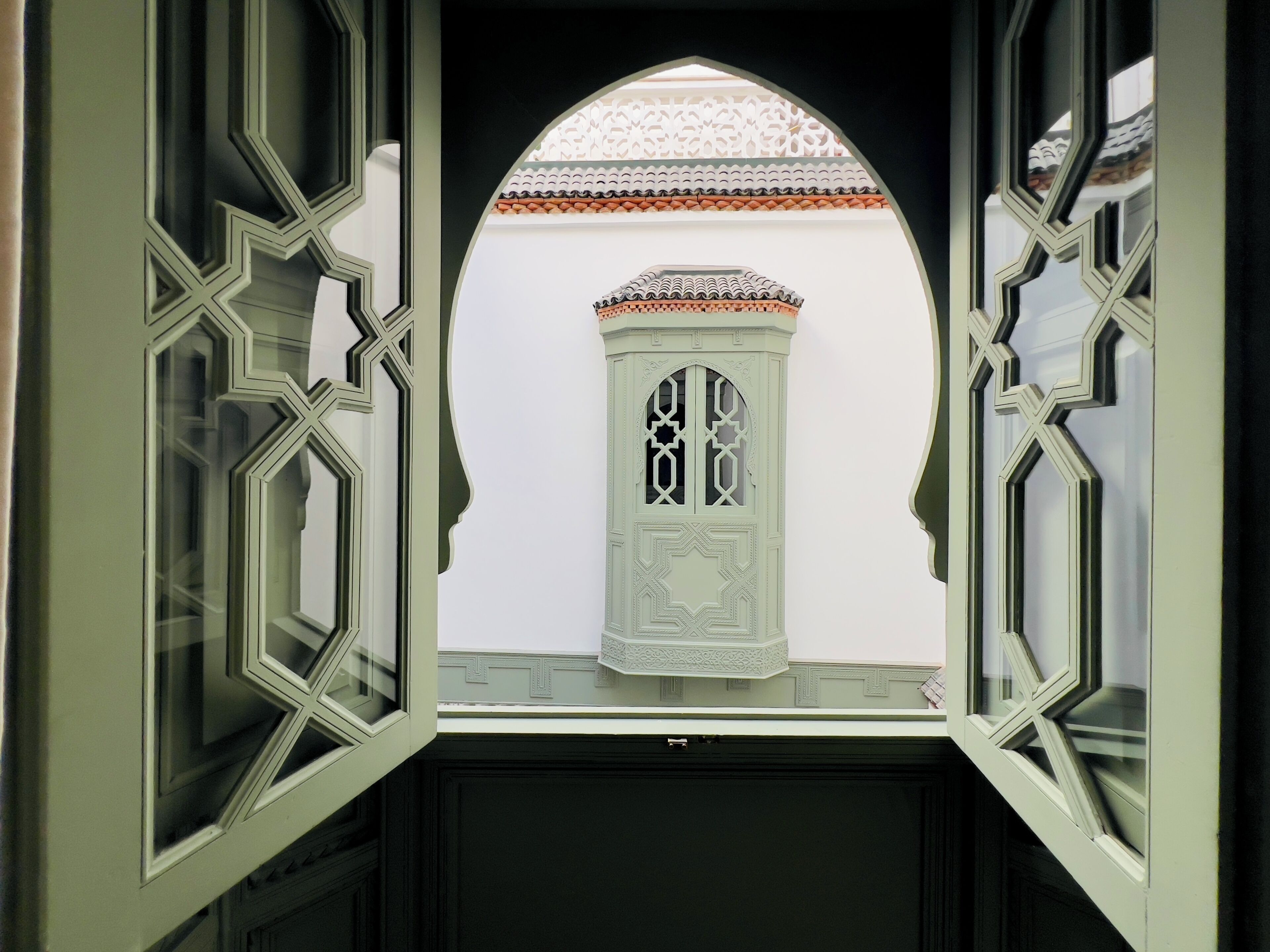 A view through an ornate window with geometric patterns, leading to a white building with a decorative roof and a door with intricate designs.