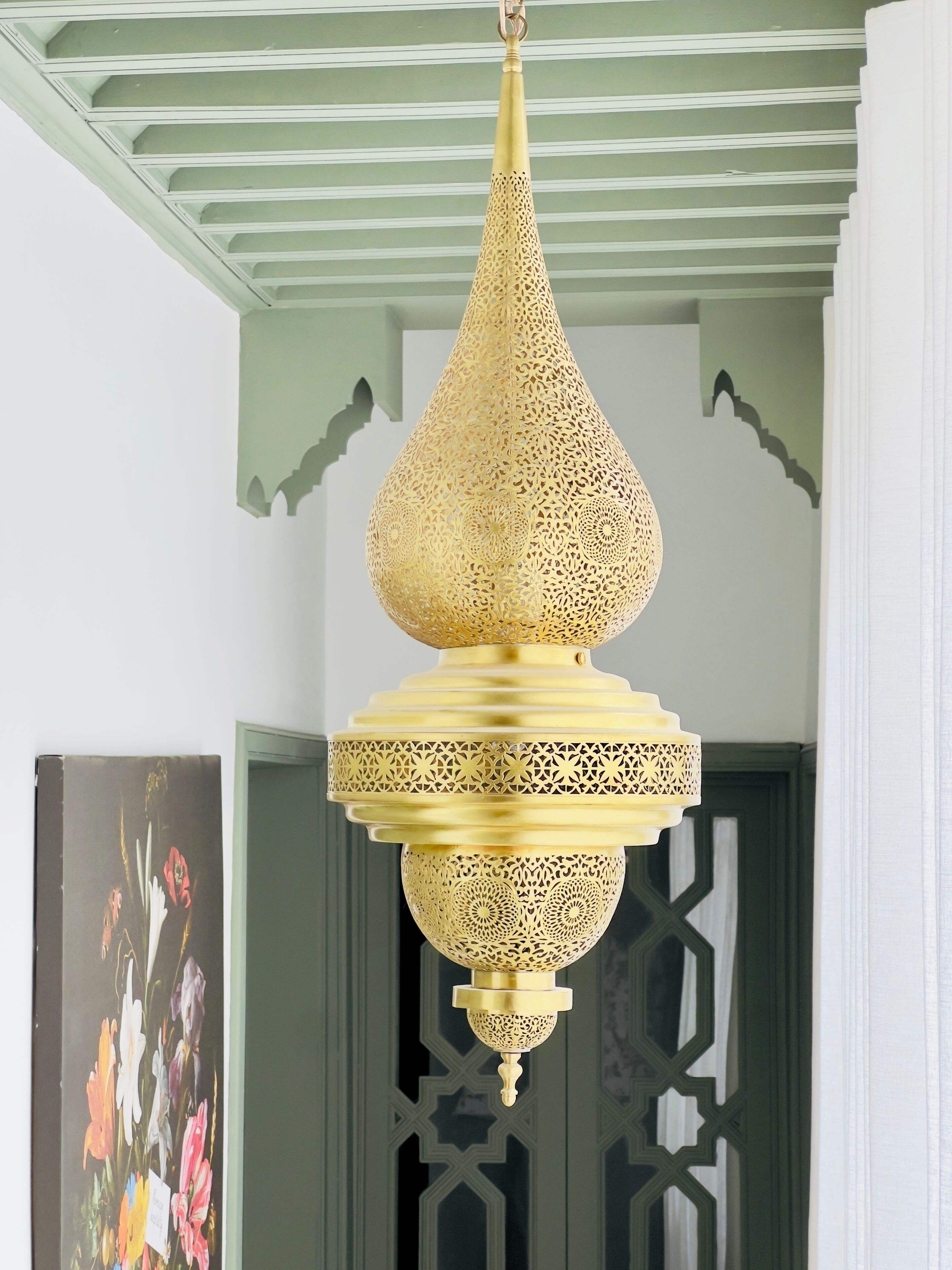 Un lampadario d'oro pendente con decorazioni elaborate, una porta verde con motivi geometrici e un quadro floreale appeso al muro.