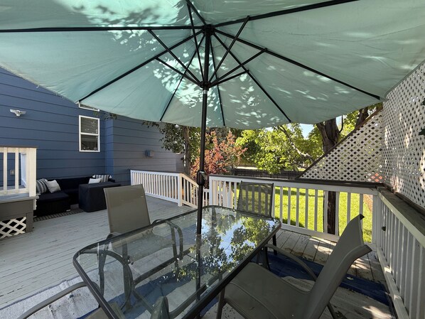 A glass-top table with chairs under a large umbrella, surrounded by a wooden deck with a white railing, and a blue house in the background.