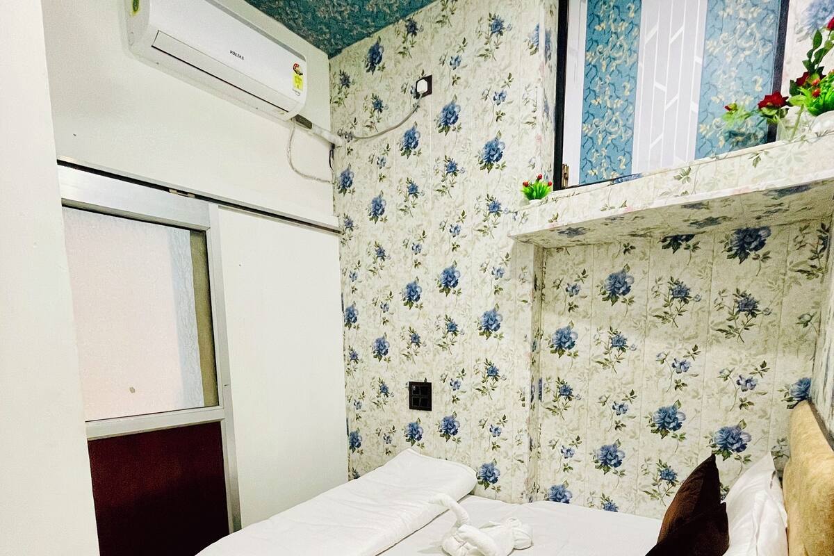 A small, single-bed room with a floral wallpaper, a white bedspread, and a brown suitcase on the floor.