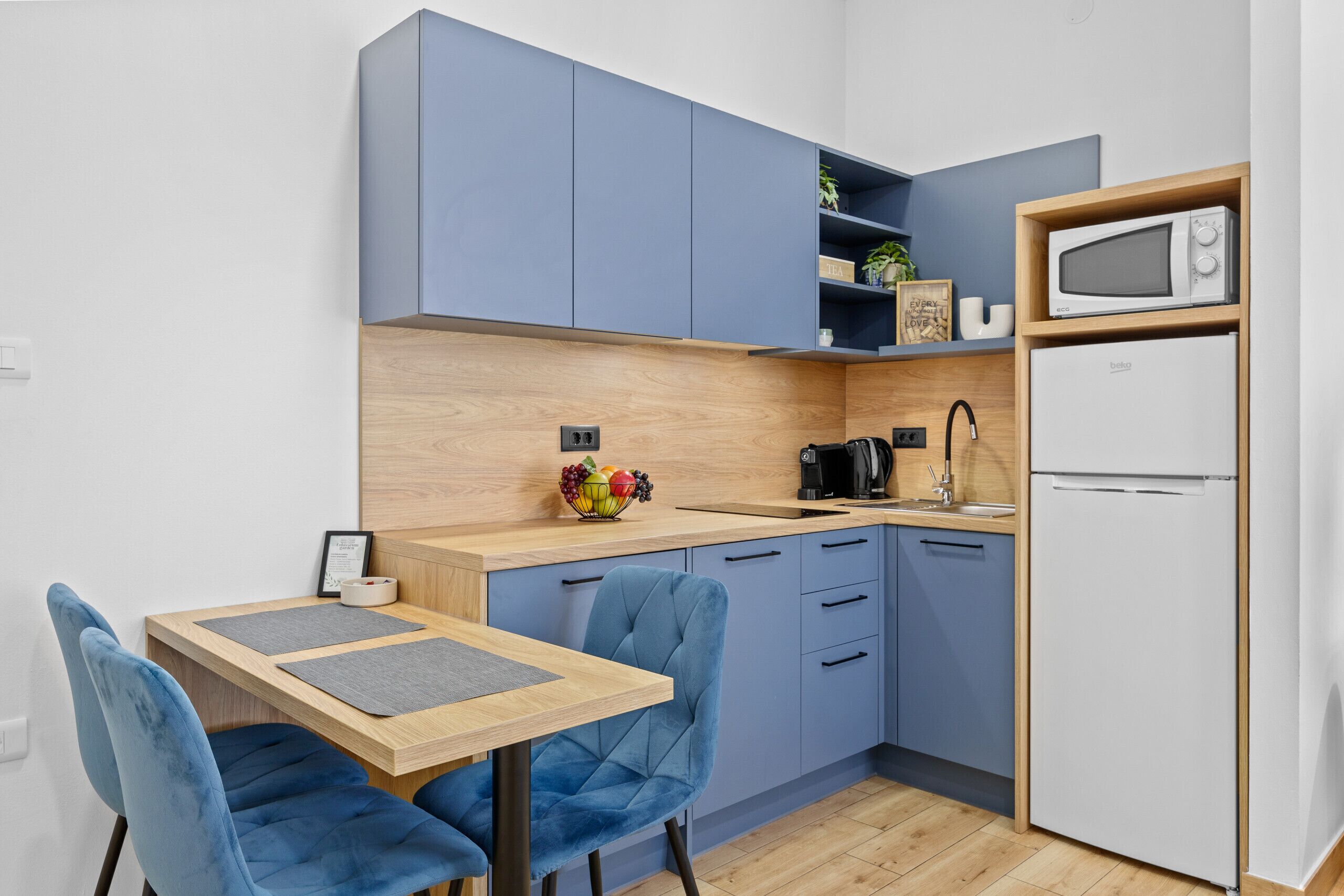 A compact kitchen with blue cabinets, a microwave, a refrigerator, a sink, and a wooden countertop. There are two blue chairs and a small table with a placemat.