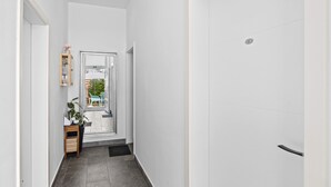 A narrow hallway with a door on the right, a small table with a plant on the left, and a view of the outdoors through a glass door.