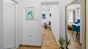 A hallway with a wooden floor, white walls, and a mirror reflecting a dining area with blue chairs and a yellow tablecloth.