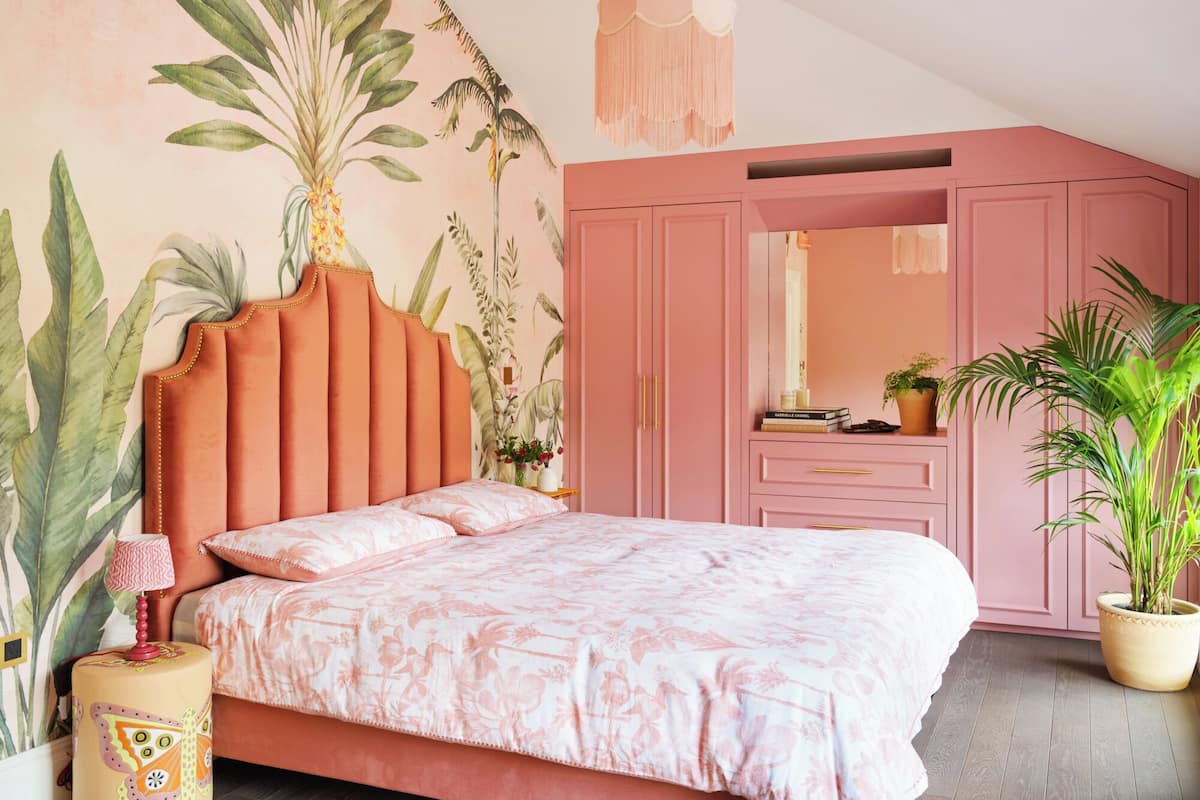 A bedroom with a pink theme, featuring a bed with a floral bedspread, a large mirror above the dresser, and a potted plant on the floor.