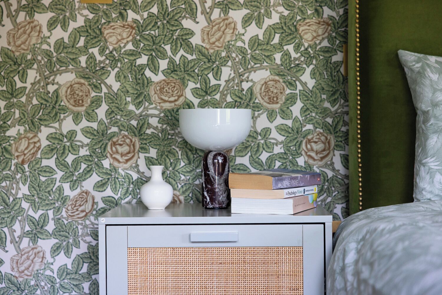 A room with a floral wallpaper, a white bedside table with a lamp, books, and a small white vase.