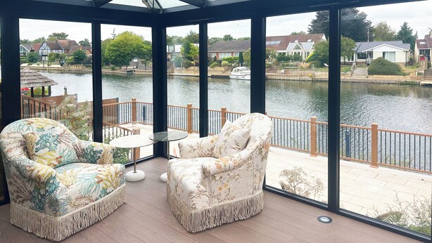 A conservatory with two floral armchairs and a small table, overlooking a calm water body and residential area. - Thames Mansion - River View (Windsor)