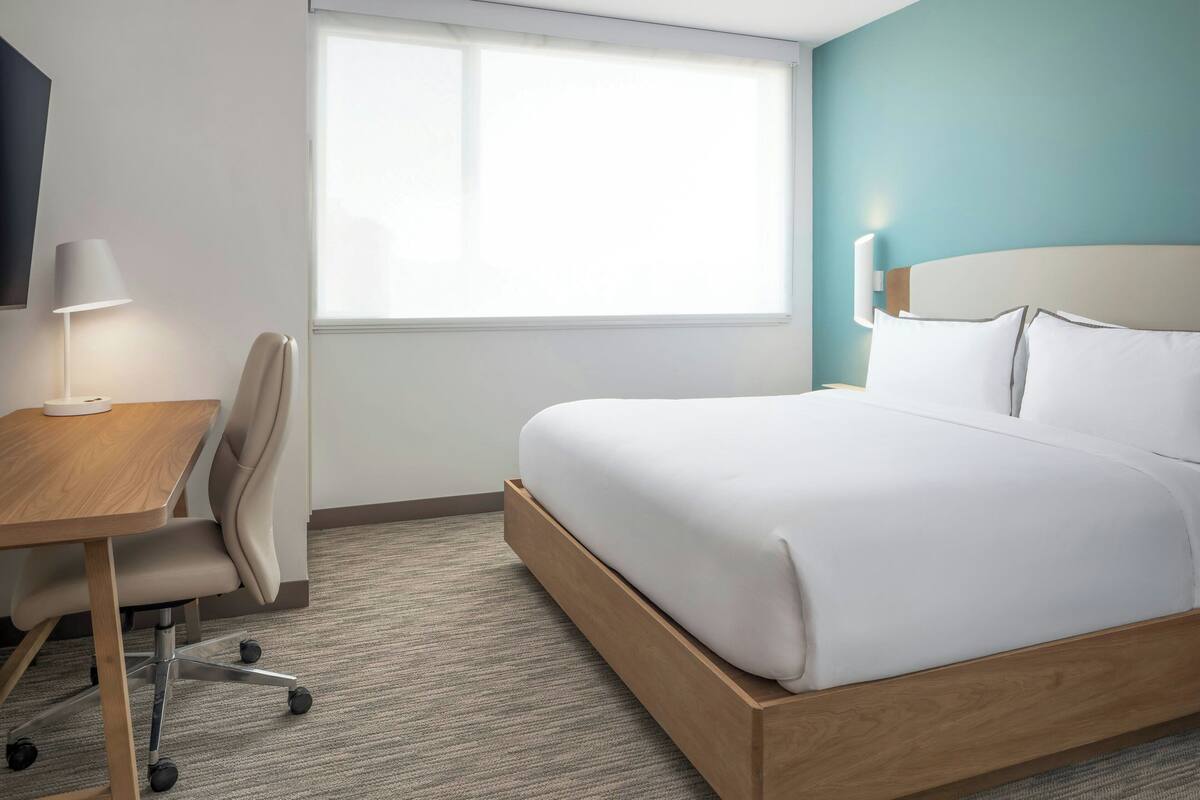 A hotel room with a bed, a desk with a chair, a lamp, and a window.