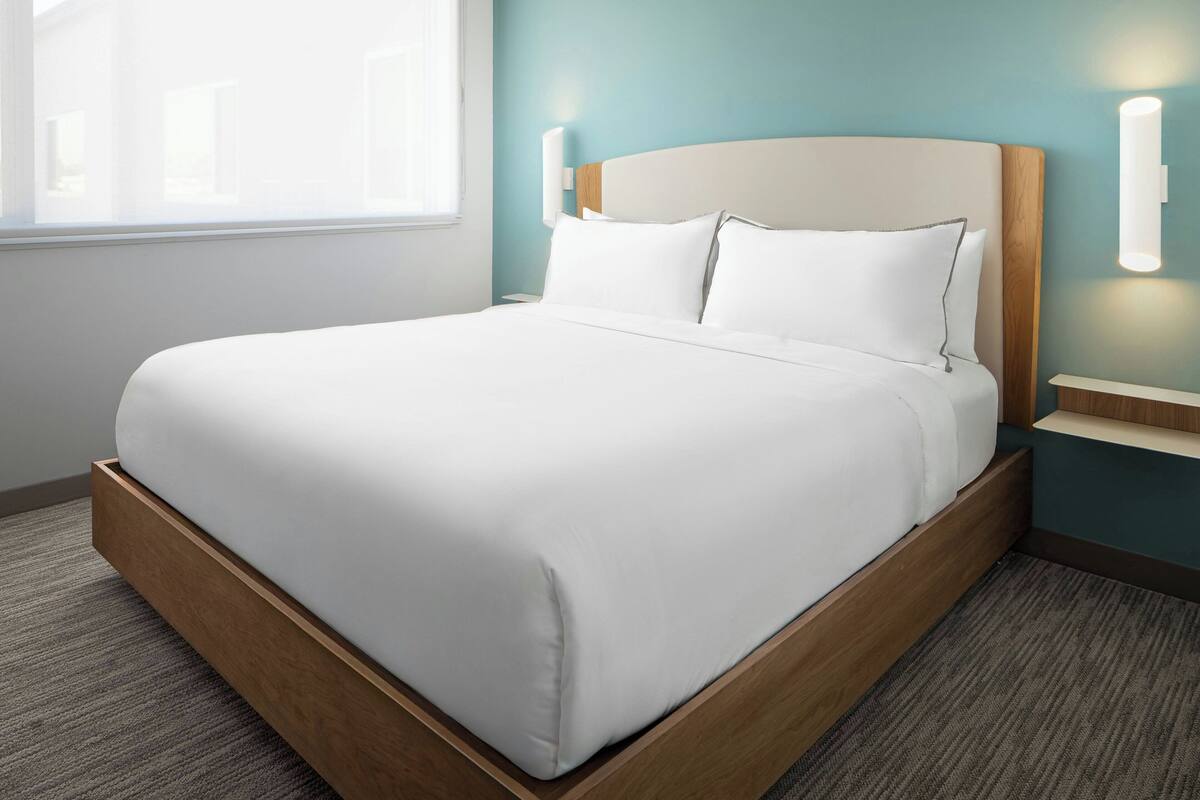 A neatly made bed in a hotel room with a wooden headboard, a nightstand, and a wall-mounted light fixture.