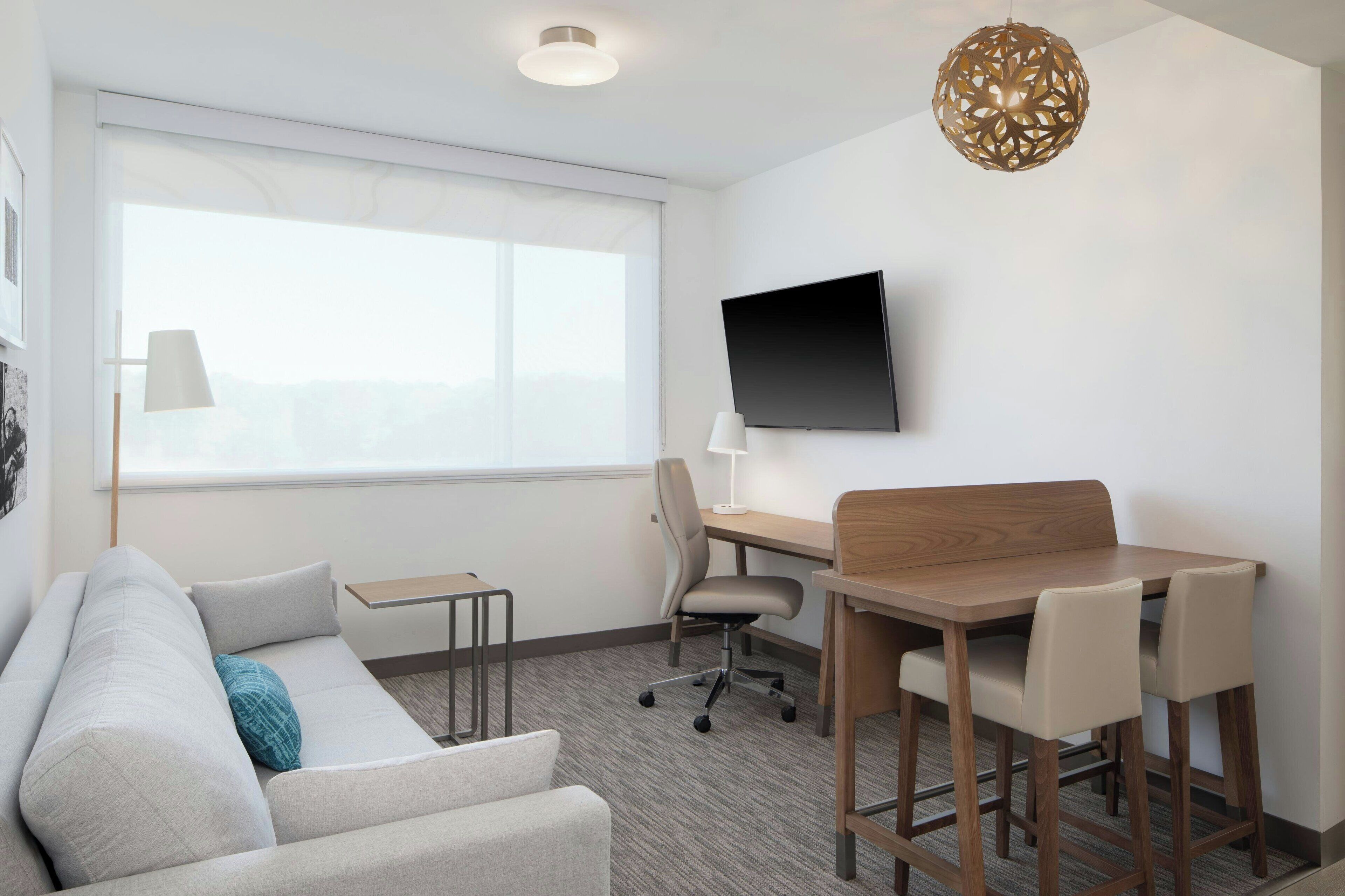 A modern hotel room with a sofa, a desk with a chair, a TV on the wall, and a large window.