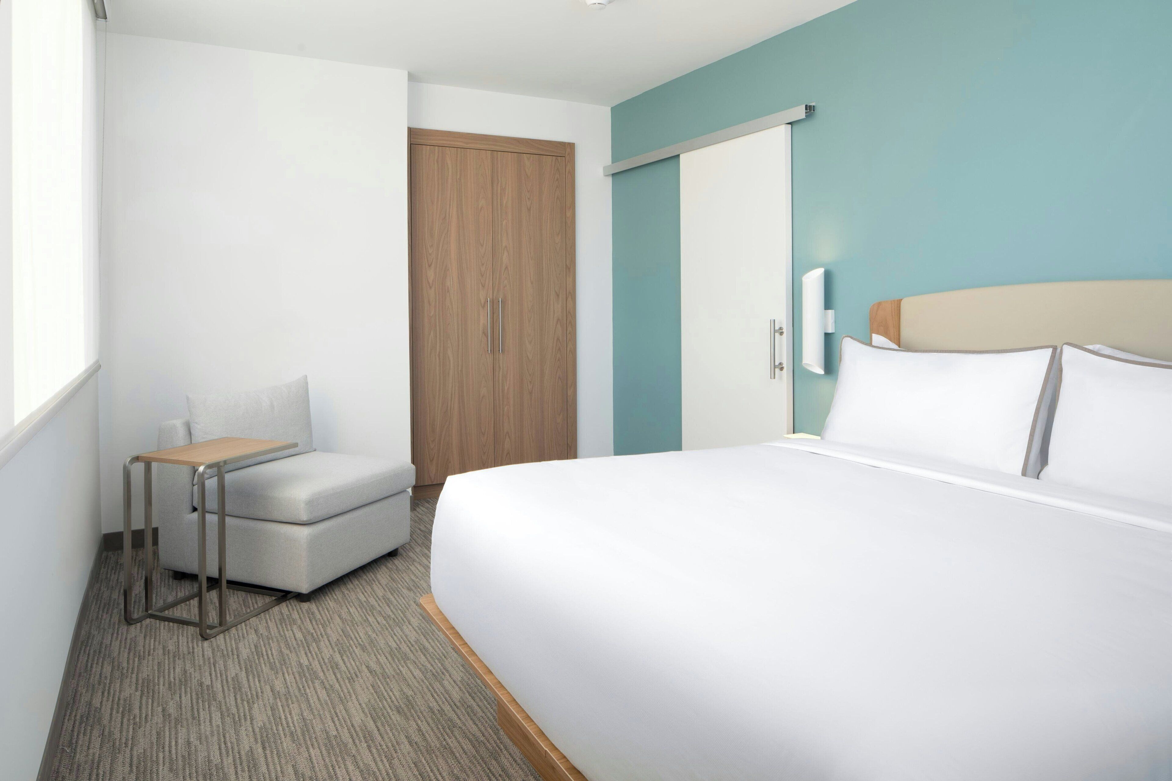 Una habitación de hotel con cama, una mesita, una silla, un armario y una ventana.