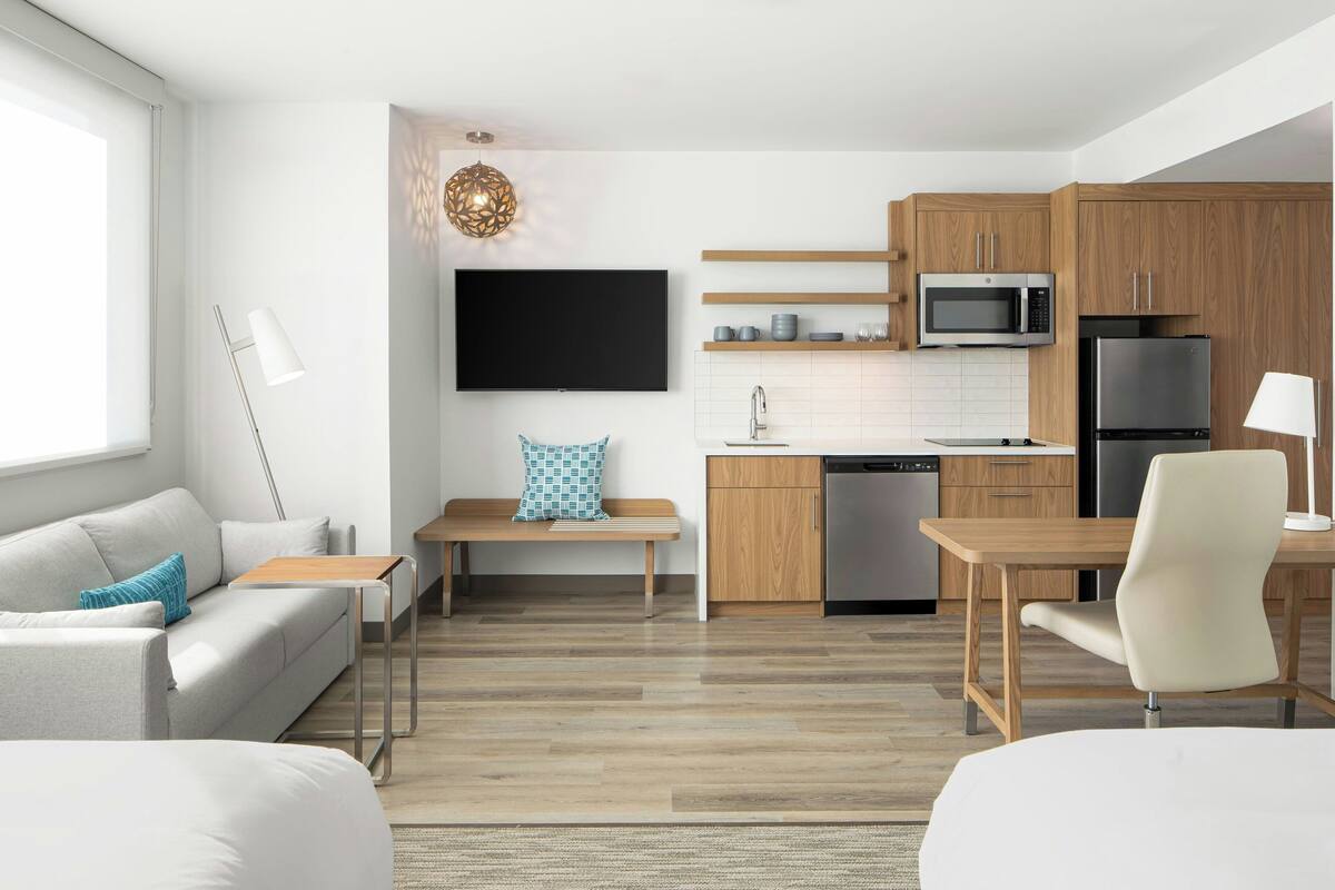 A modern kitchen with wooden cabinets, a microwave, and a refrigerator. There is a dining table with chairs, a bench, and a flat-screen TV mounted on the wall.