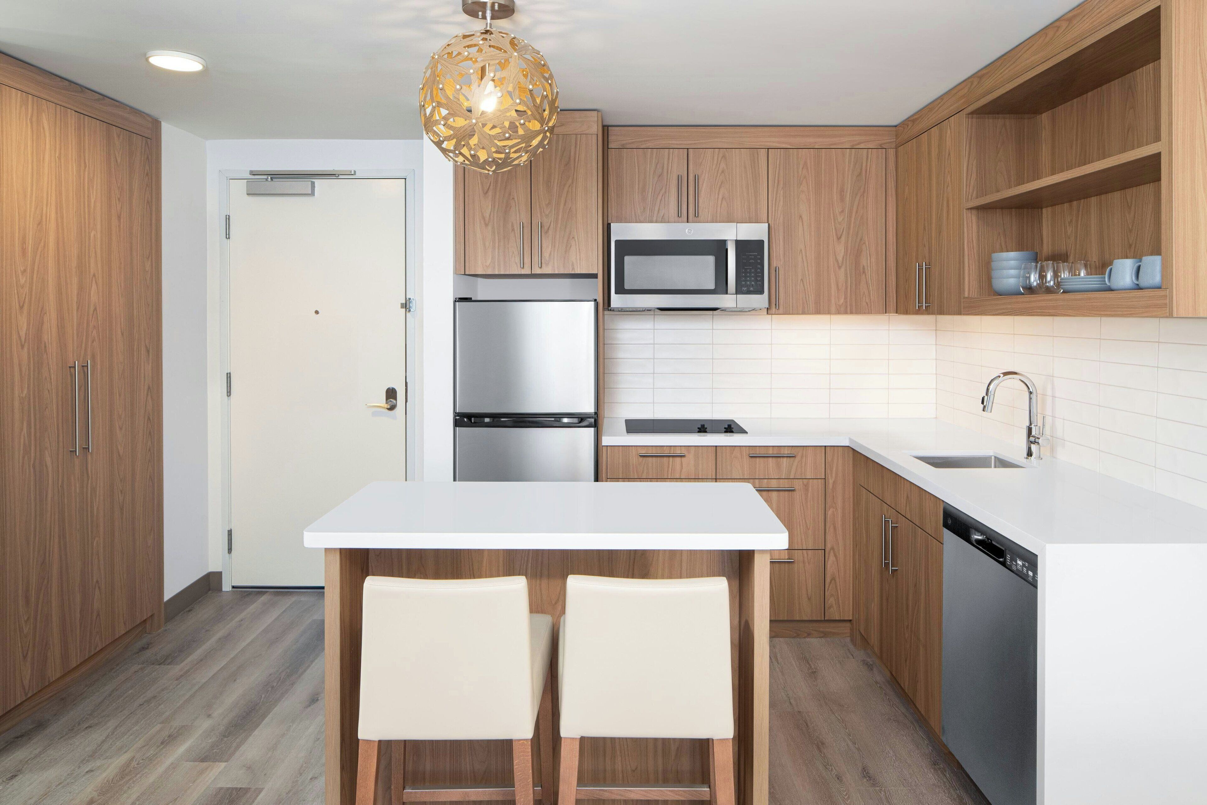 Una cocina moderna con armarios de madera, una encimera blanca, un refrigerador de acero inoxidable, un microondas y un lavavajillas. Hay una puerta blanca y una lámpara colgante sobre la isla de la cocina.