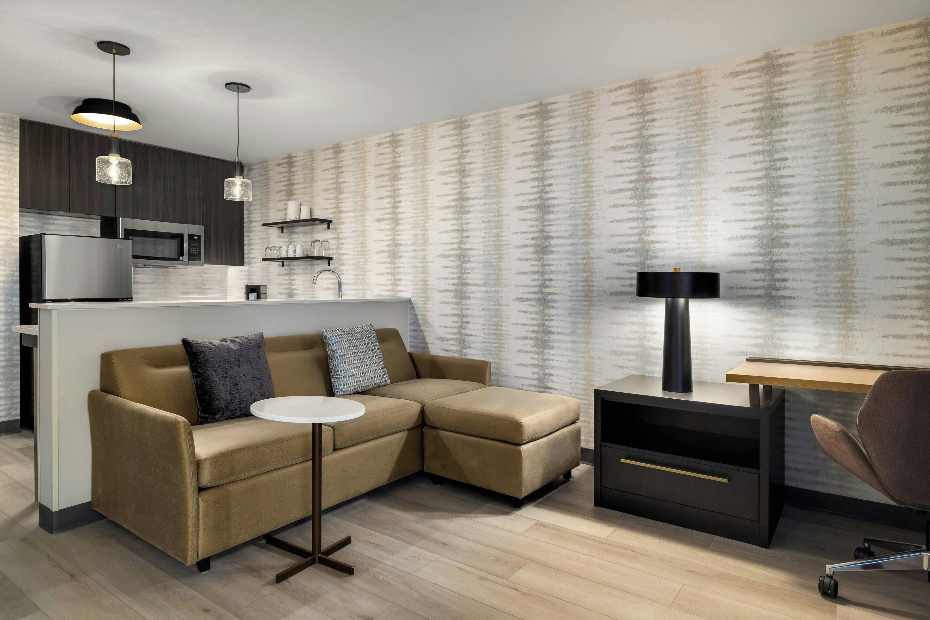 A modern living space with a sofa, a small round table, a desk, and a lamp. There is a kitchen area with a microwave and a refrigerator.