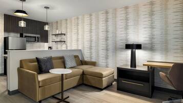 A modern living space with a sofa, a small round table, a desk, and a lamp. There is a kitchen area with a microwave and a refrigerator.