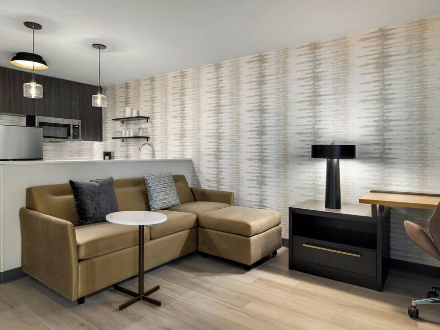 A modern living space with a sofa, a small round table, a desk, and a lamp. There is a kitchen area with a microwave and a refrigerator.