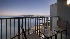 A balcony with a view of a calm body of water and a distant pier. There is a chair and a table on the balcony.