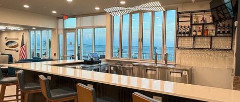 A modern bar area with a long counter, bar stools, and a view of the ocean through large windows.