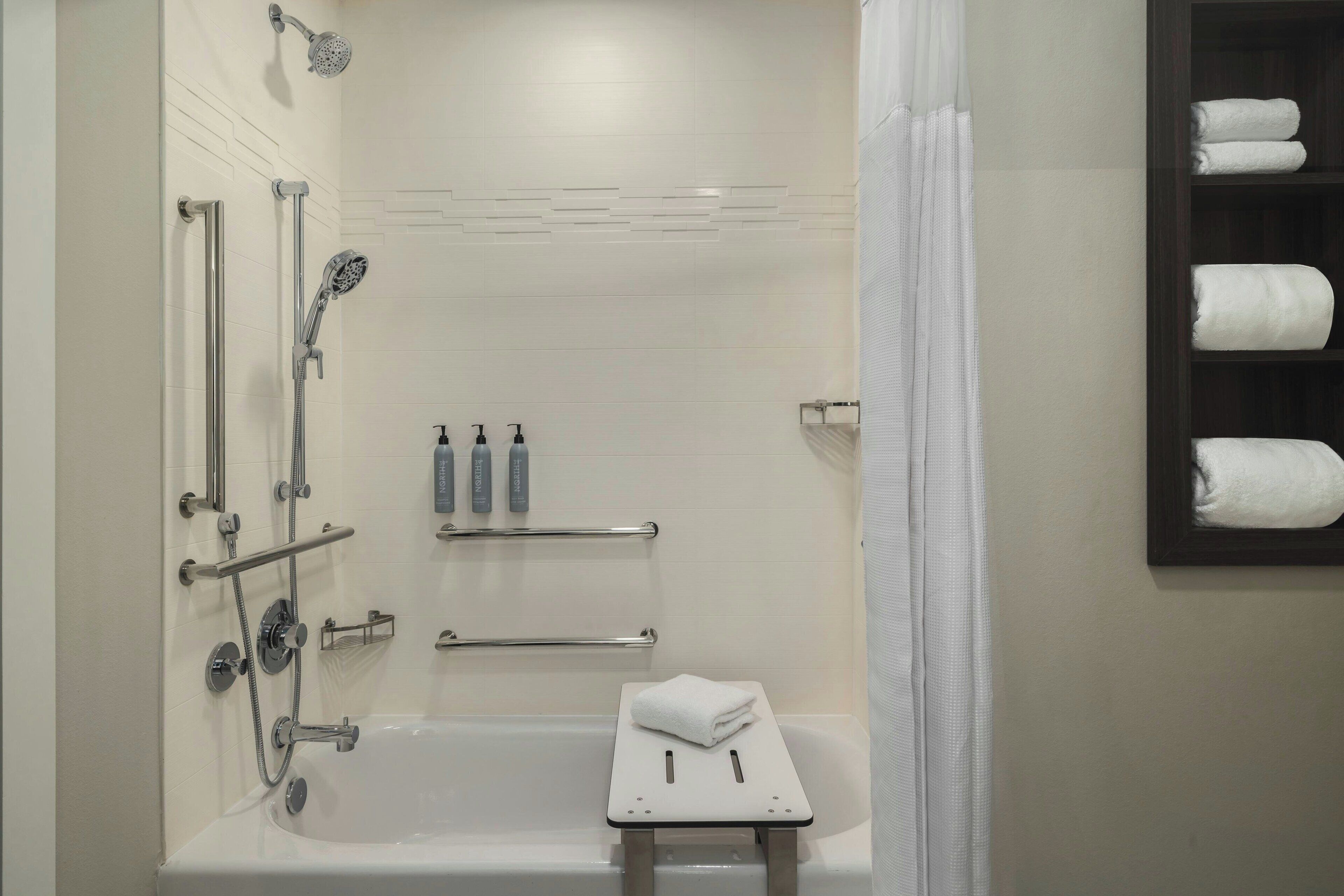 Ein modernes Badezimmer mit Badewanne, Dusche und Haltegriffen. Handtücher und Pflegeprodukte befinden sich an der Wand.