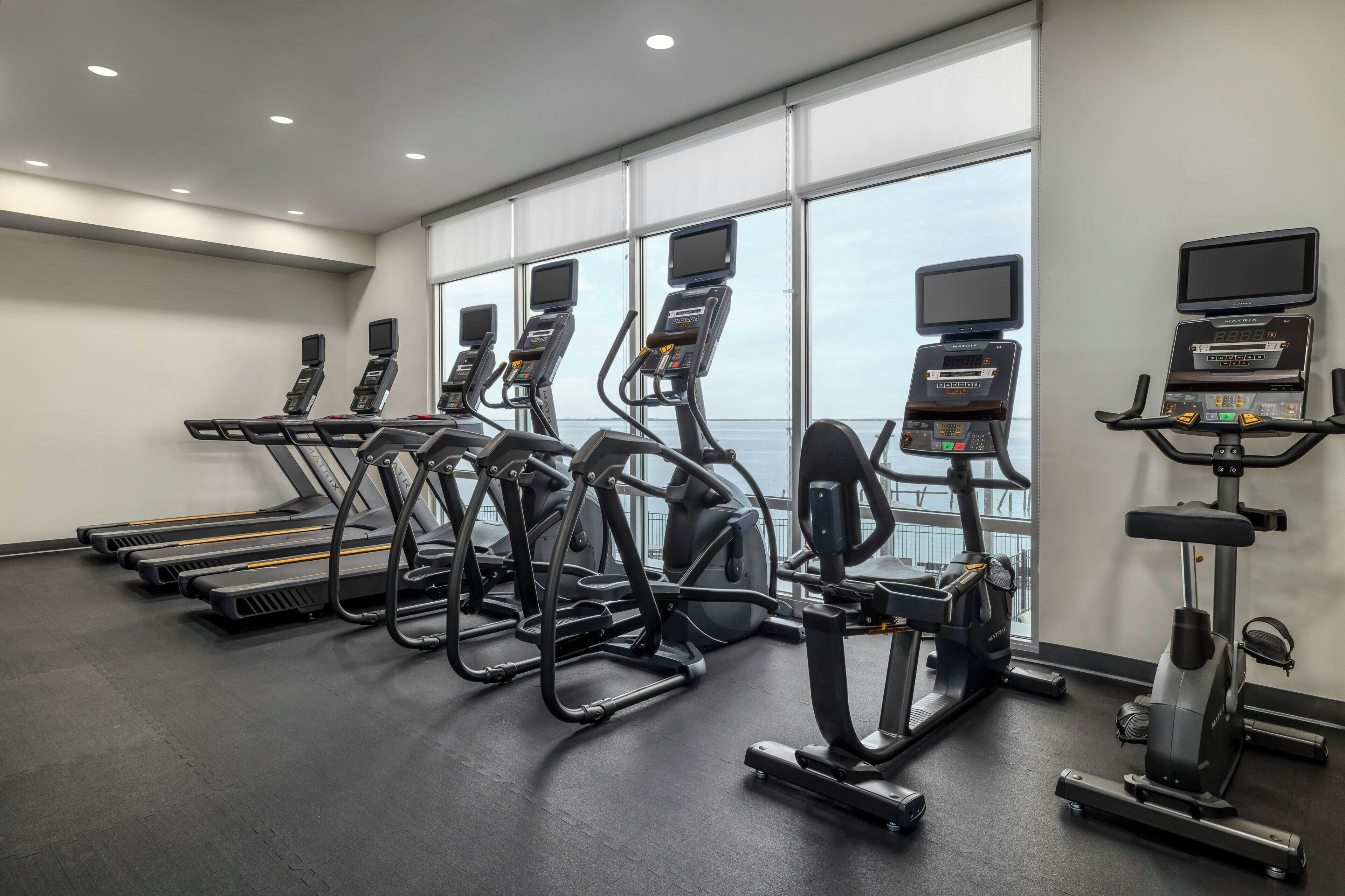 A gym with treadmills, elliptical machines, and a rowing machine, featuring large windows and a view of the sky.