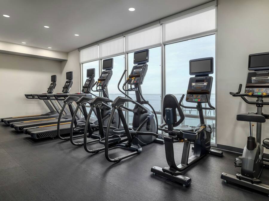 Et fitnesscenter med løbebånd, crosstrainere og romaskine, med store vinduer og udsigt til himlen.