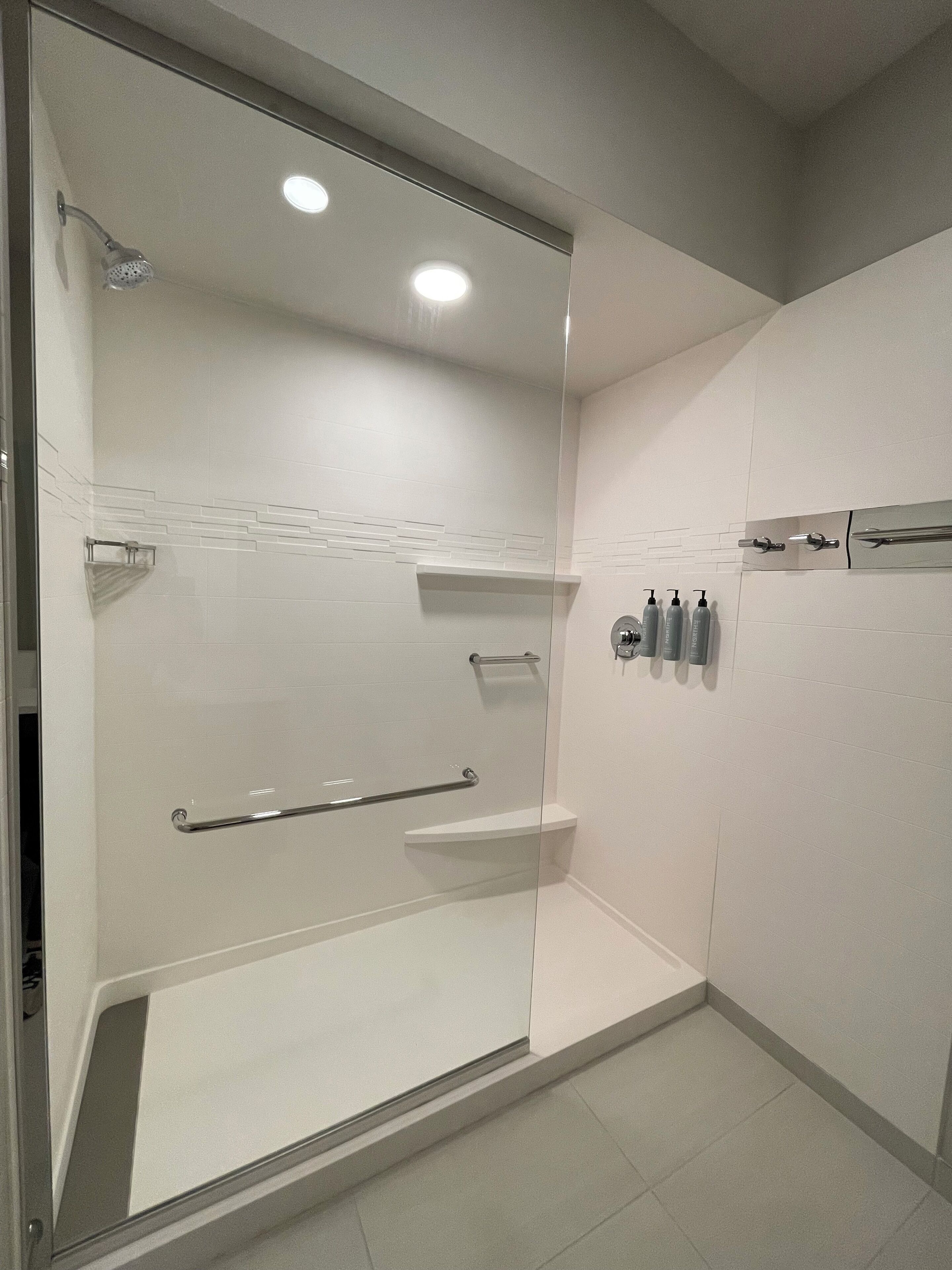 A modern bathroom with a glass shower enclosure, a handrail, and a shelf with toiletries.