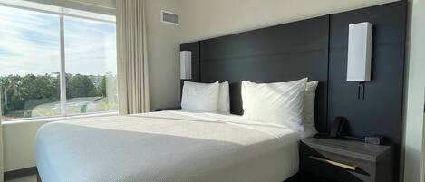A modern hotel room with a large bed, a nightstand, a wall-mounted lamp, and a window with curtains.
