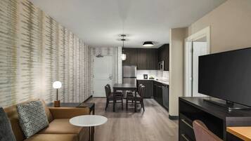 A modern living room with a sofa, a small round table, and a TV. The room has a kitchen in the background with a refrigerator, microwave, and cabinets.
