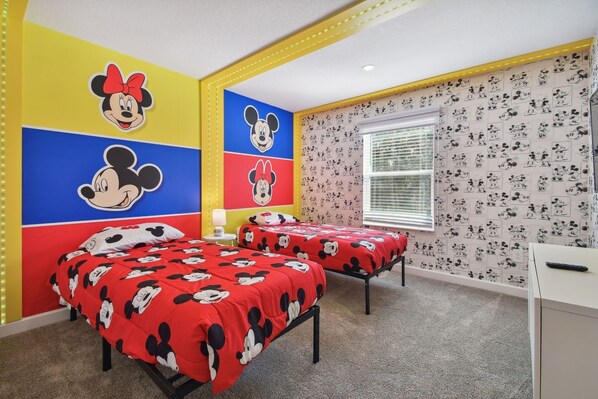 A room with two beds, each covered with red sheets featuring Mickey Mouse prints, a white desk, and walls decorated with Mickey Mouse images.