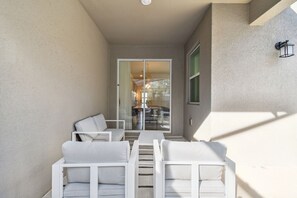 A patio area with white furniture, a glass door leading to an interior space, and a wall-mounted light fixture.