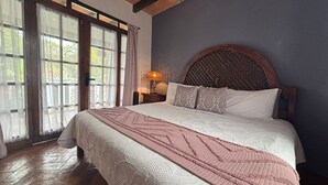 A bedroom with a large bed, a wooden headboard, a nightstand with a lamp, and a view through a window with curtains.