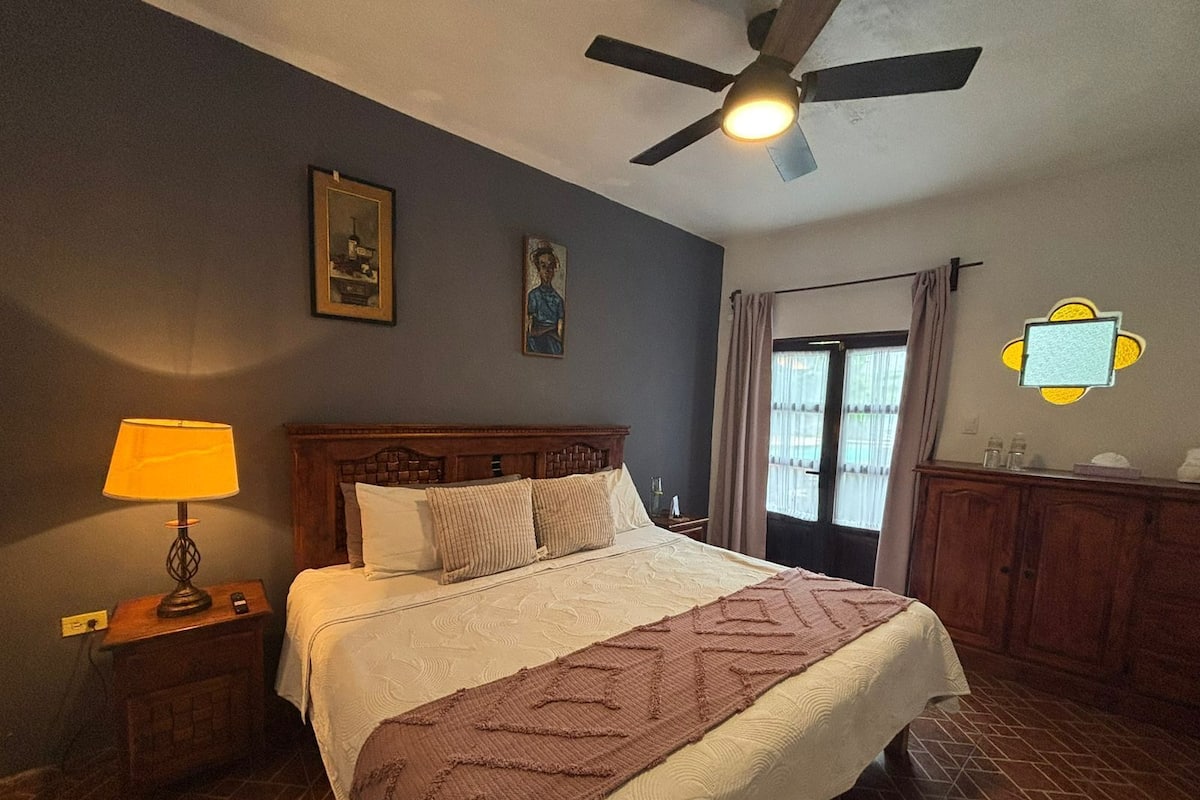 Quarto com cama, mesa de cabeceira, luminária, ventilador de teto e janela com cortinas.