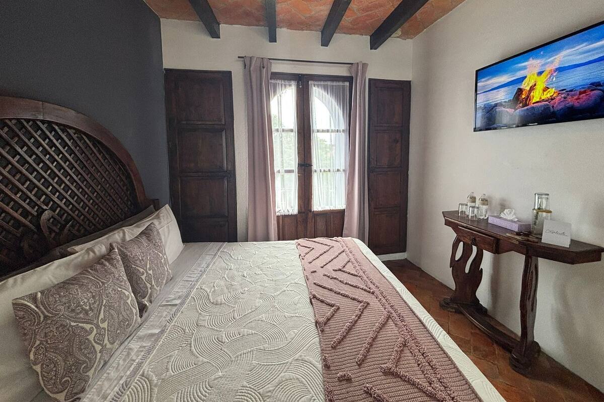 Quarto com cama, cabeceira de madeira, mesa de cabeceira, quadro e janela com cortinas.
