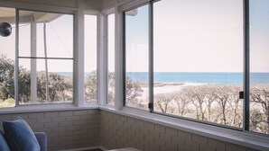 Een kamer met grote ramen met uitzicht op een strand, een bakstenen muur en een kleine tafel met een theepot en kopjes.