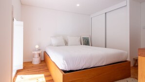 A bedroom with a wooden bed, white bedding, a small round nightstand with a lamp, and a framed picture on the wall.